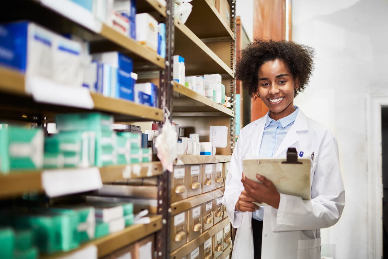 Druggist at medicine storage room at pharmacy Confident African female pharmacist standing by a medicine shelf holding a clipboard.